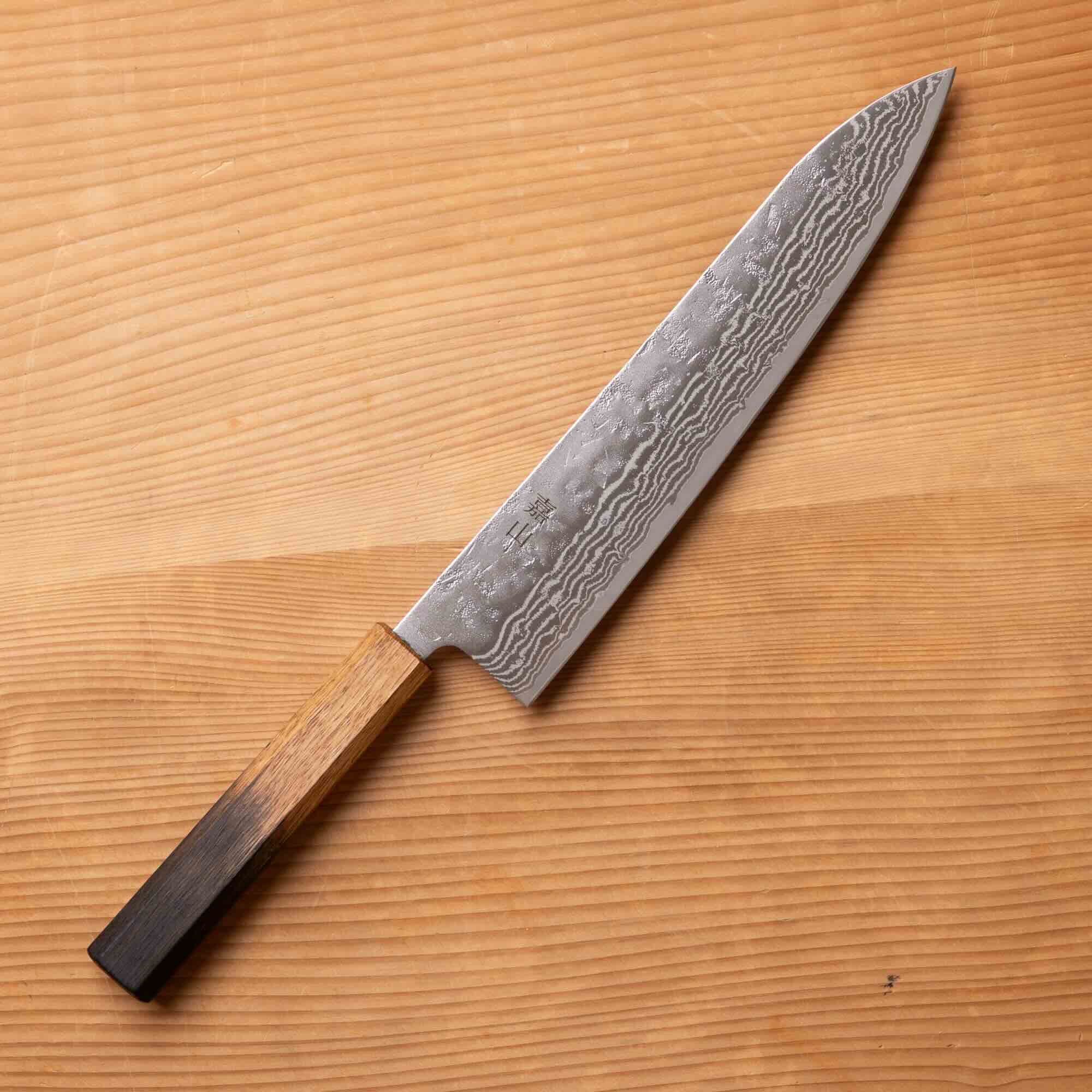 A Japanese-style kitchen knife with a patterned Damascus steel blade and a dark wood handle, placed diagonally on a light brown wooden surface.