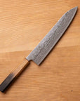 A Japanese-style kitchen knife with a patterned Damascus steel blade and a dark wood handle, placed diagonally on a light brown wooden surface.