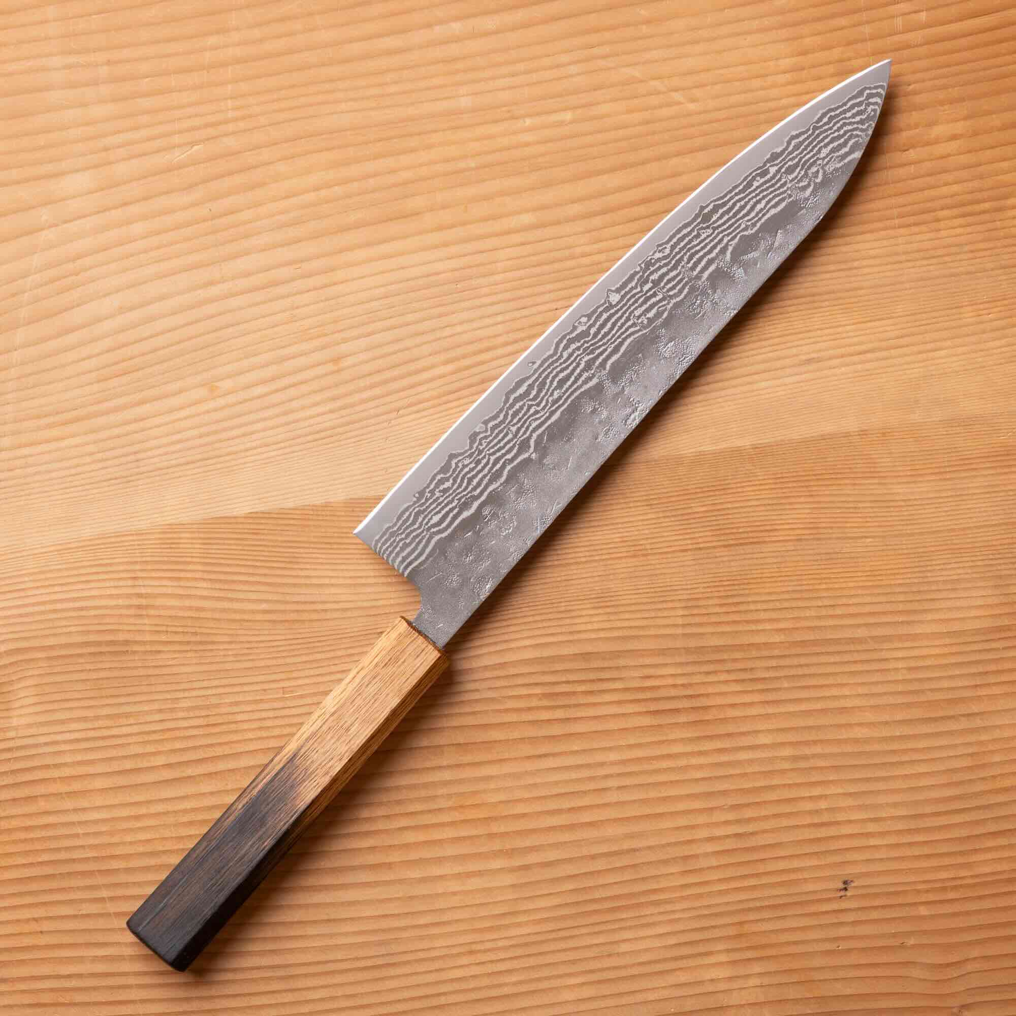 A Japanese-style kitchen knife with a Damascus steel blade and a wooden handle, placed diagonally on a light wood grain surface.