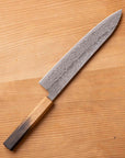 A Japanese-style kitchen knife with a Damascus steel blade and a wooden handle, placed diagonally on a light wood grain surface.
